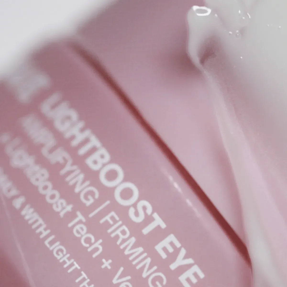 Close-up of a pink label for Solawaves LightBoost Eye Cream, featuring Eye Amplifying/Firming enhanced by LightBoost technology. A glossy, translucent cream is artfully smeared on the surface.